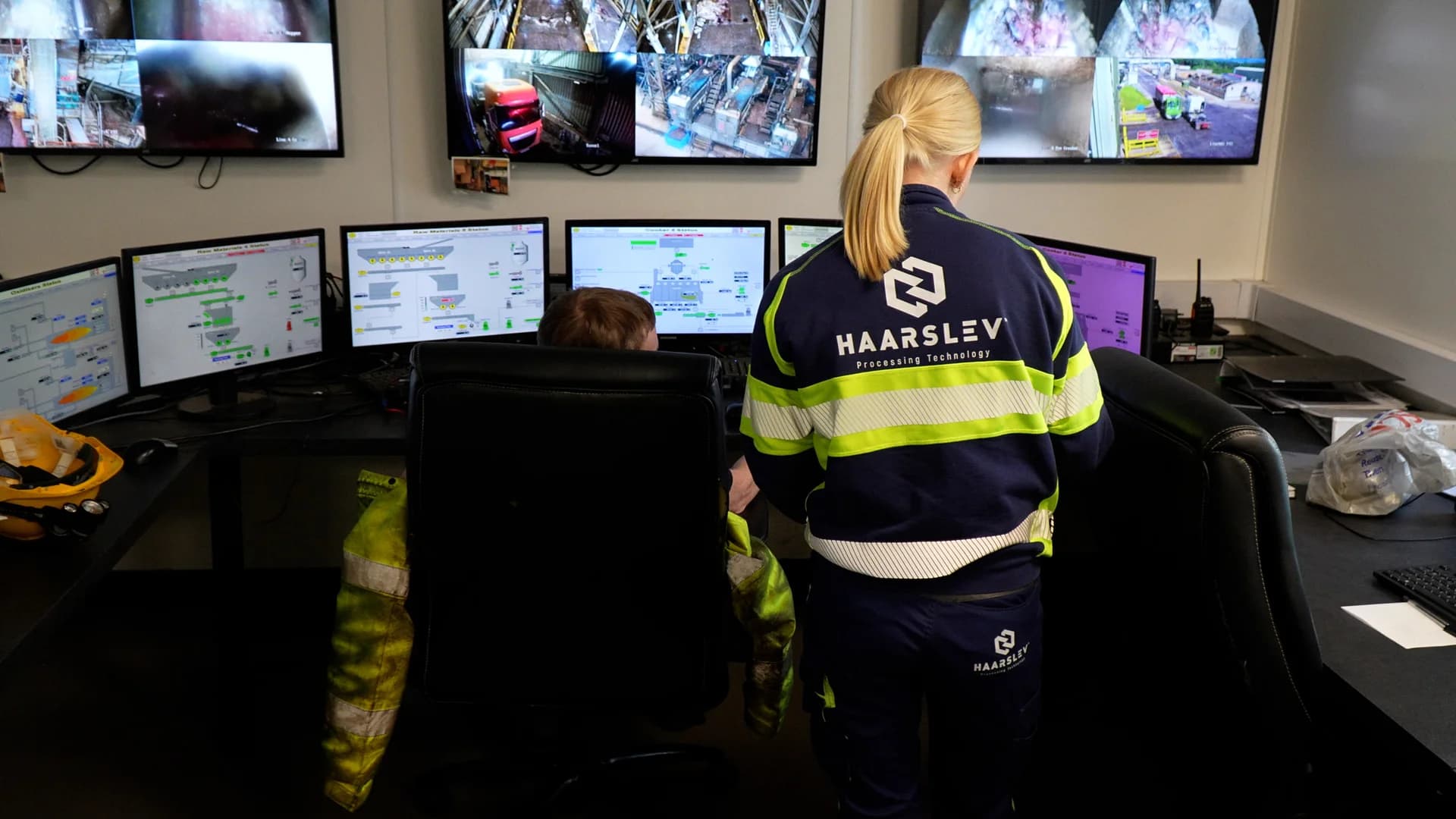 A Haarslev Automation engineer works alongside an Advanced Proteins operator in the site's control room during CORE commissioning. The screens show live process graphics from the rendering line, with CCTV feeds of the plant mounted above.